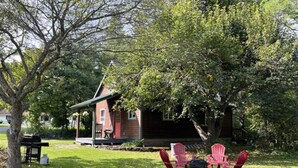 Un chalet rustique doté d’un porche, entouré d’une végétation luxuriante et agrémenté d’un espace pique-nique avec des chaises et un foyer.