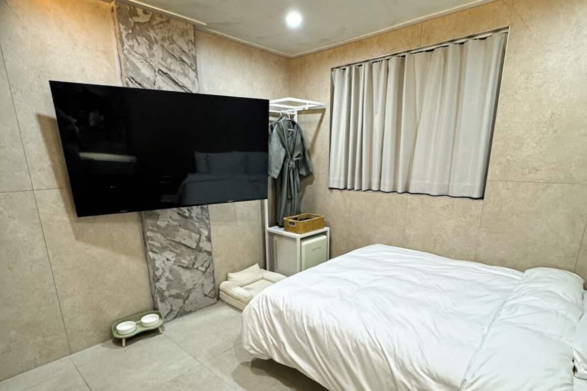 A modern bedroom with a flat-screen TV mounted on the wall, a bed with white bedding, a small bedside table, and a window with curtains.