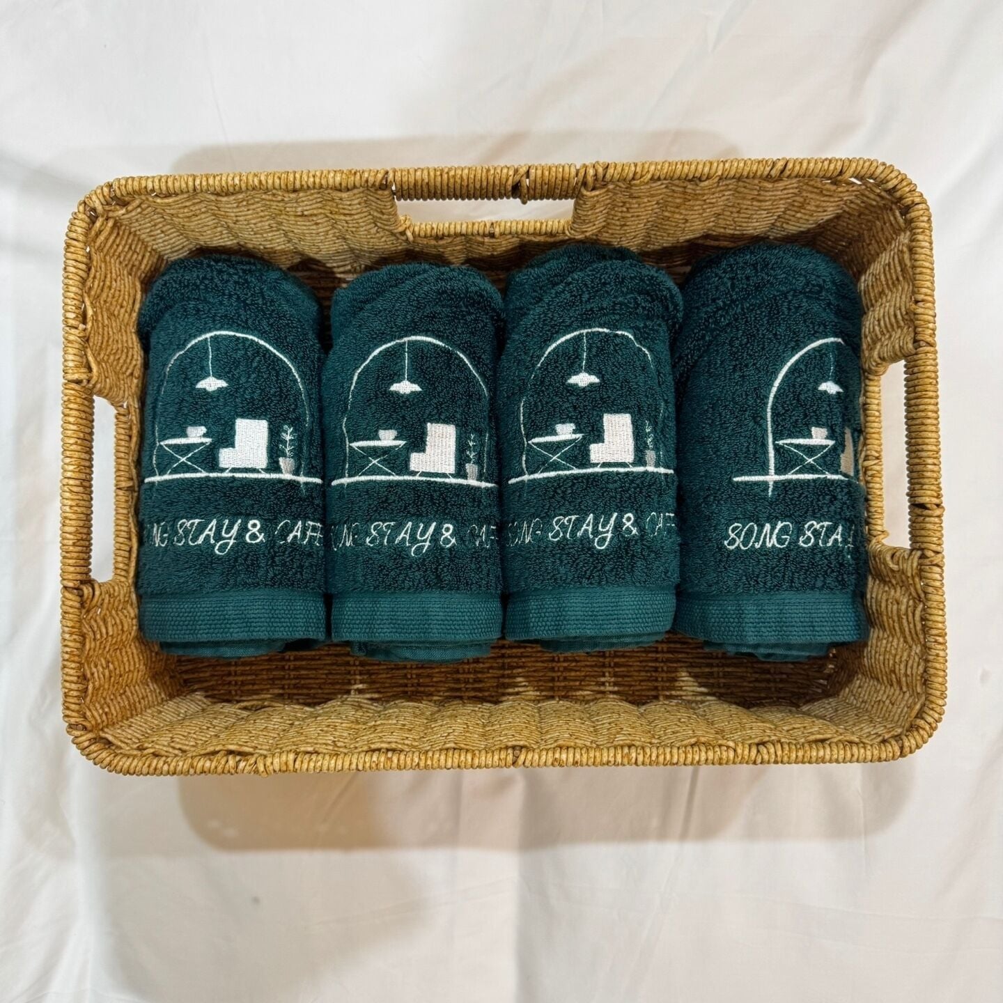 Una cesta de mimbre que contiene cuatro toallas verde oscuro con diseños y texto bordados.
