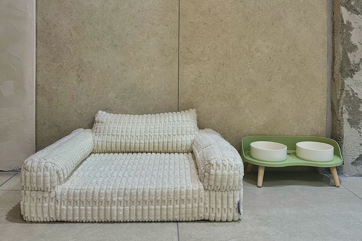 A small, textured sofa with a matching pillow against a plain wall, next to a green tray with two bowls.