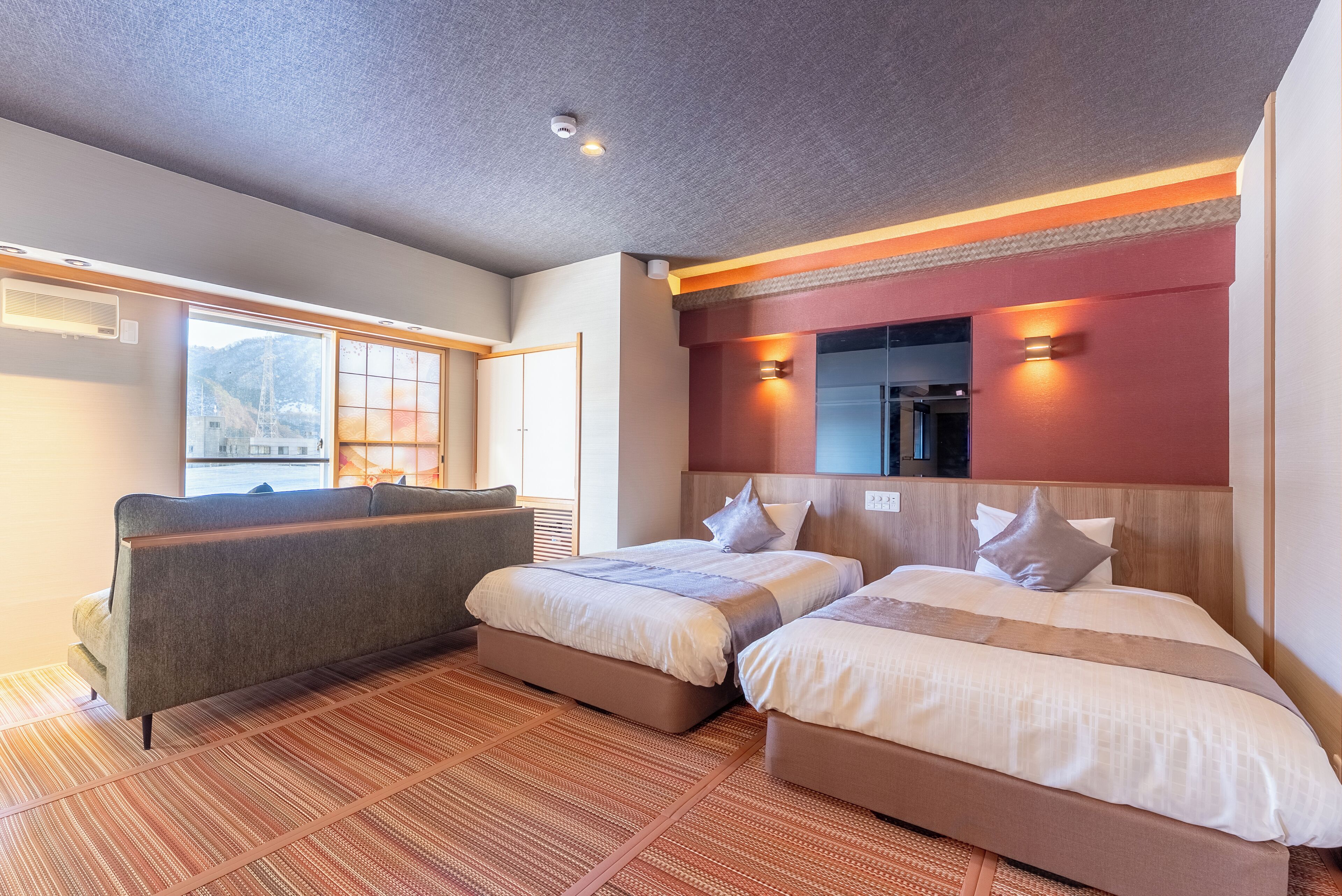 A hotel room with two beds, a sofa, a large window with a view of mountains, and a red accent wall.