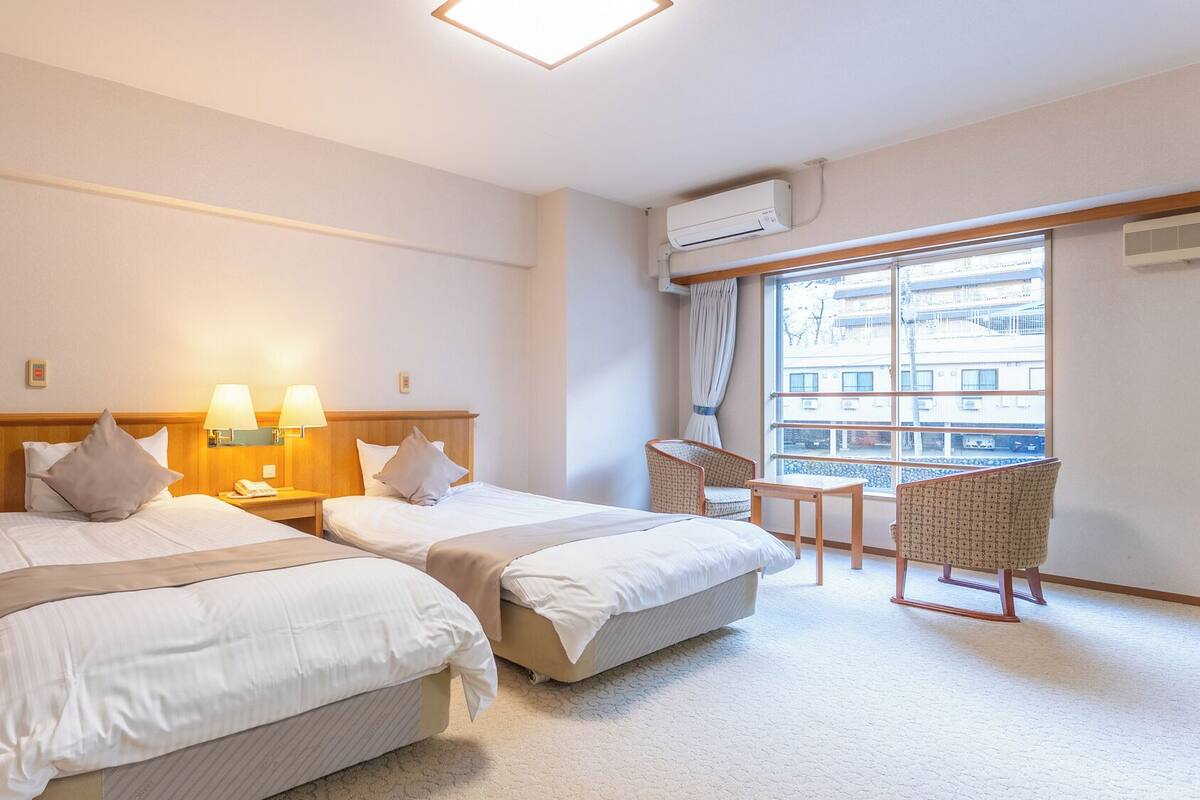 A hotel room with two beds, a small table, two chairs, and a large window with a view of buildings.