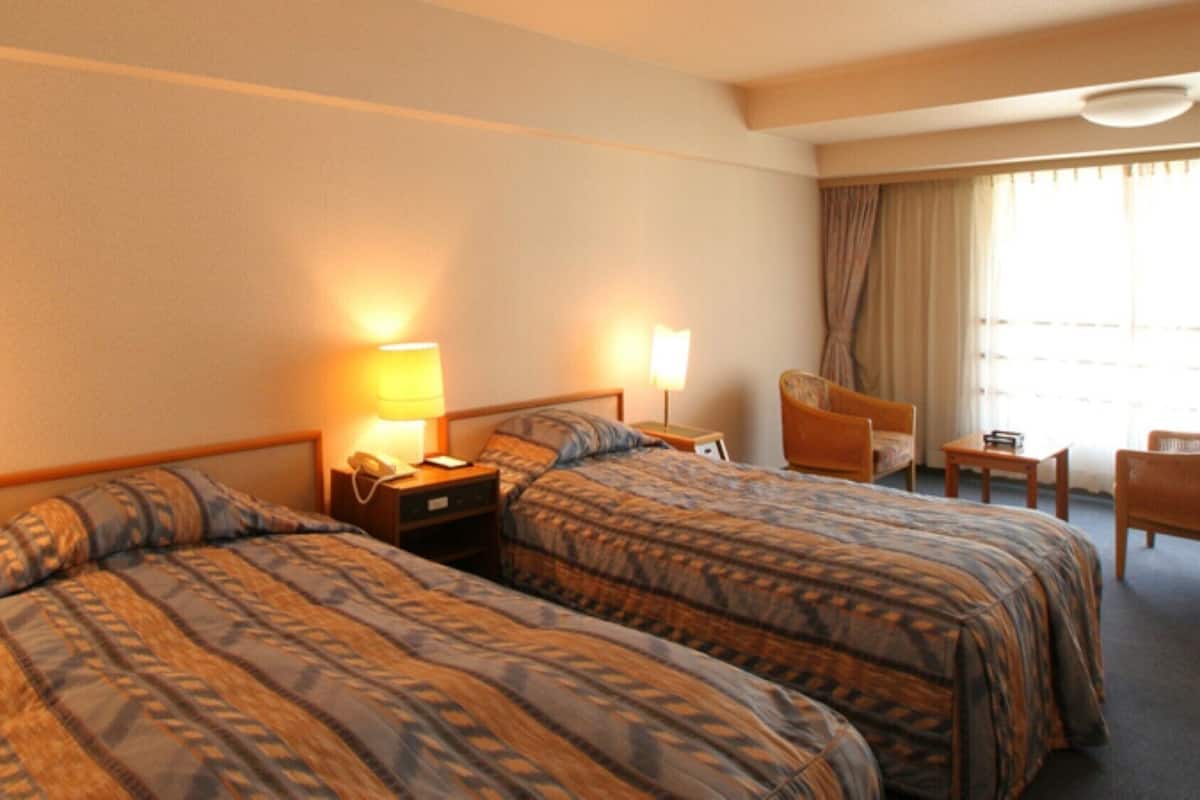 A hotel room with two beds, a chair, a small table, and a window with curtains.