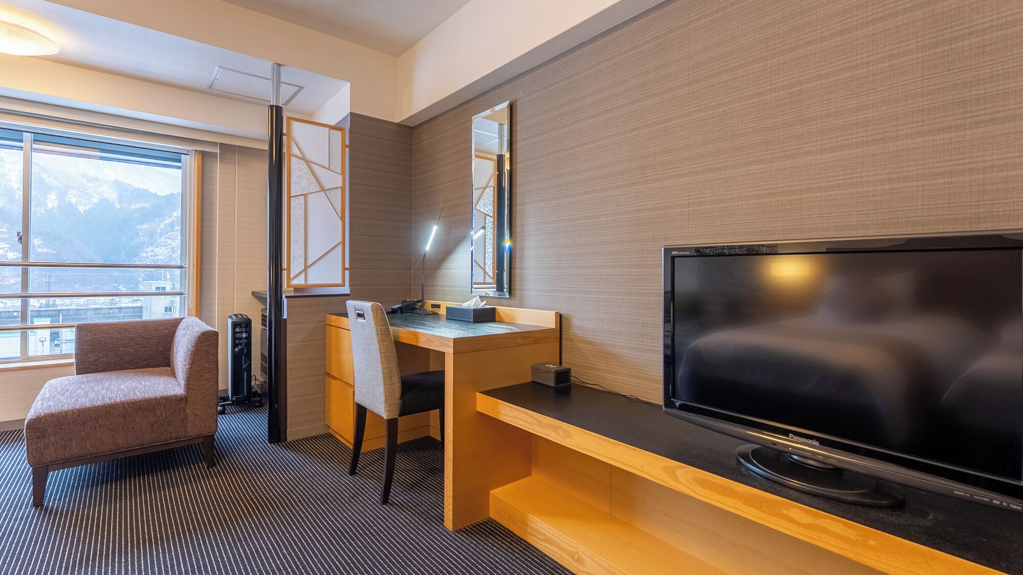 A modern hotel room with a large flat-screen TV, a wooden desk, a comfortable armchair, and a large window offering a mountain view.