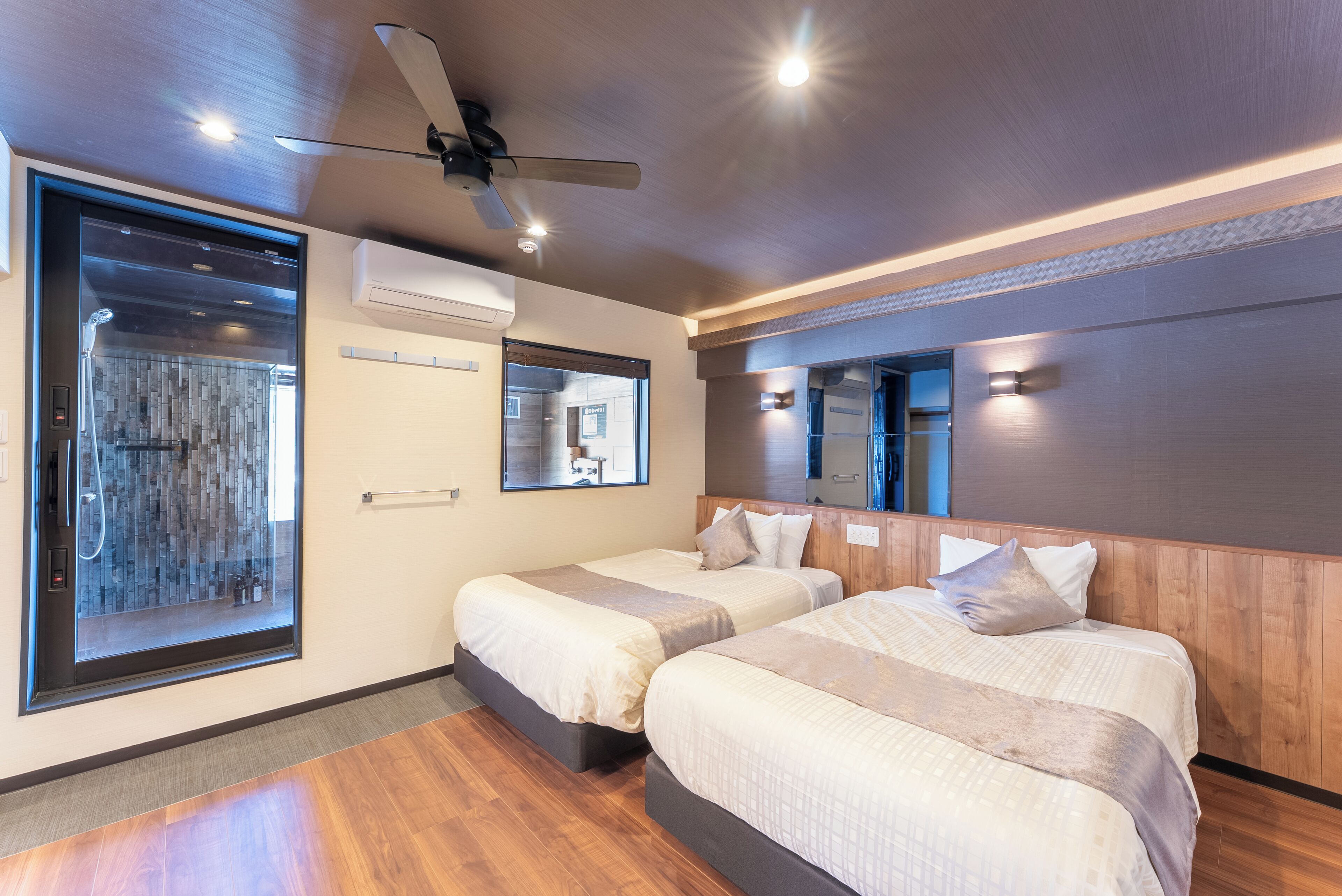 A hotel room with two beds, a ceiling fan, a large window, and a shower area.