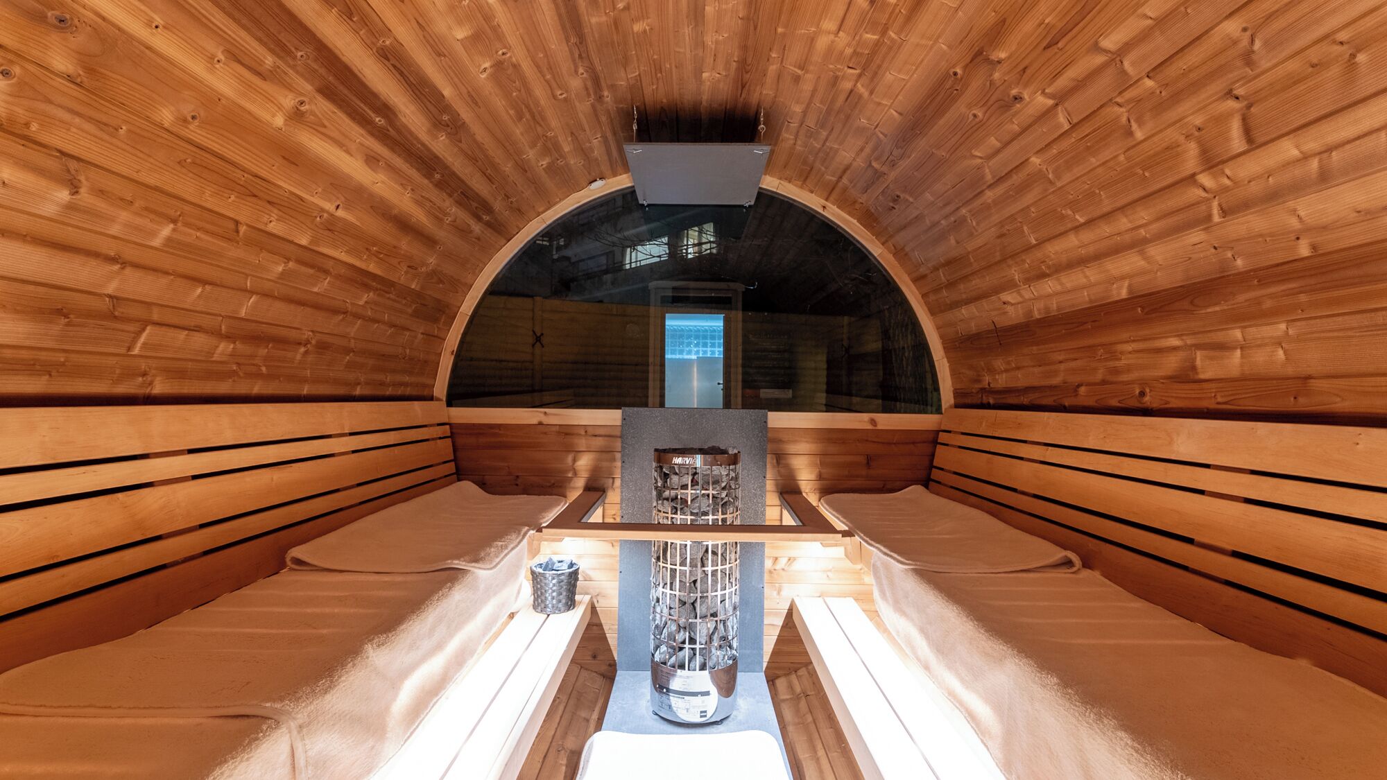 A sauna with wooden walls and benches, a central heater, and a glass door leading outside.