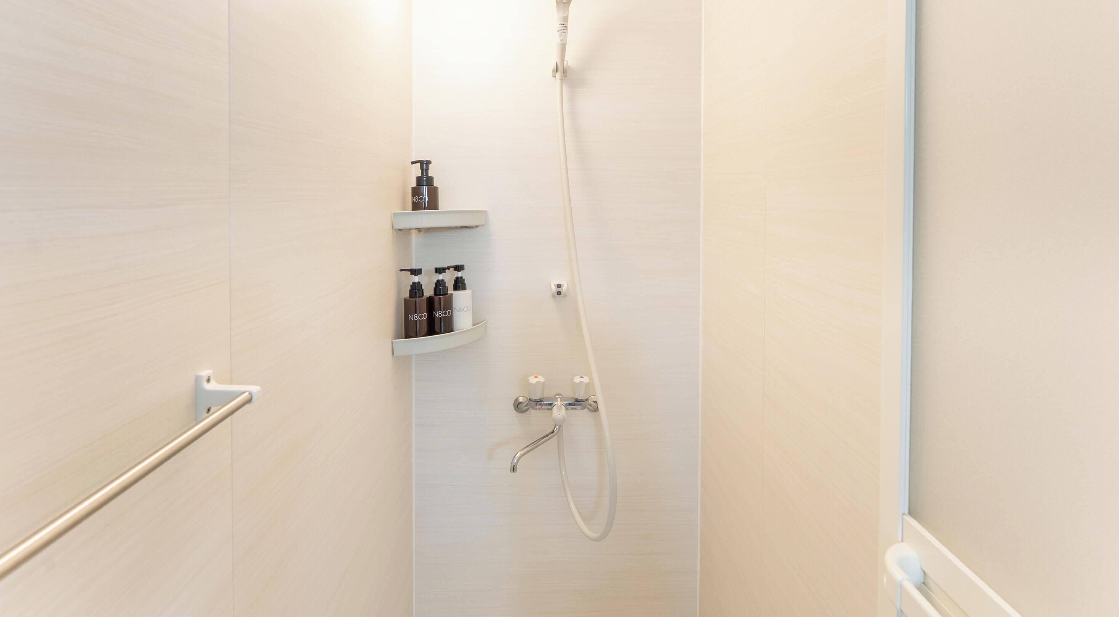 A narrow shower area with a white showerhead and handheld shower attachment, a white towel rack, and a shelf with toiletries.