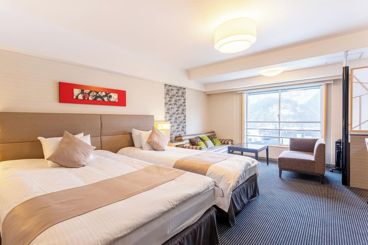 A hotel room with two beds, a seating area with a chair, a large window, and artwork on the wall.