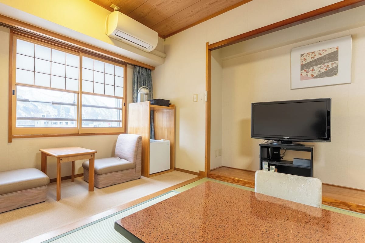 A room with a television, two cushioned seating areas, a small table, and a window with a view.