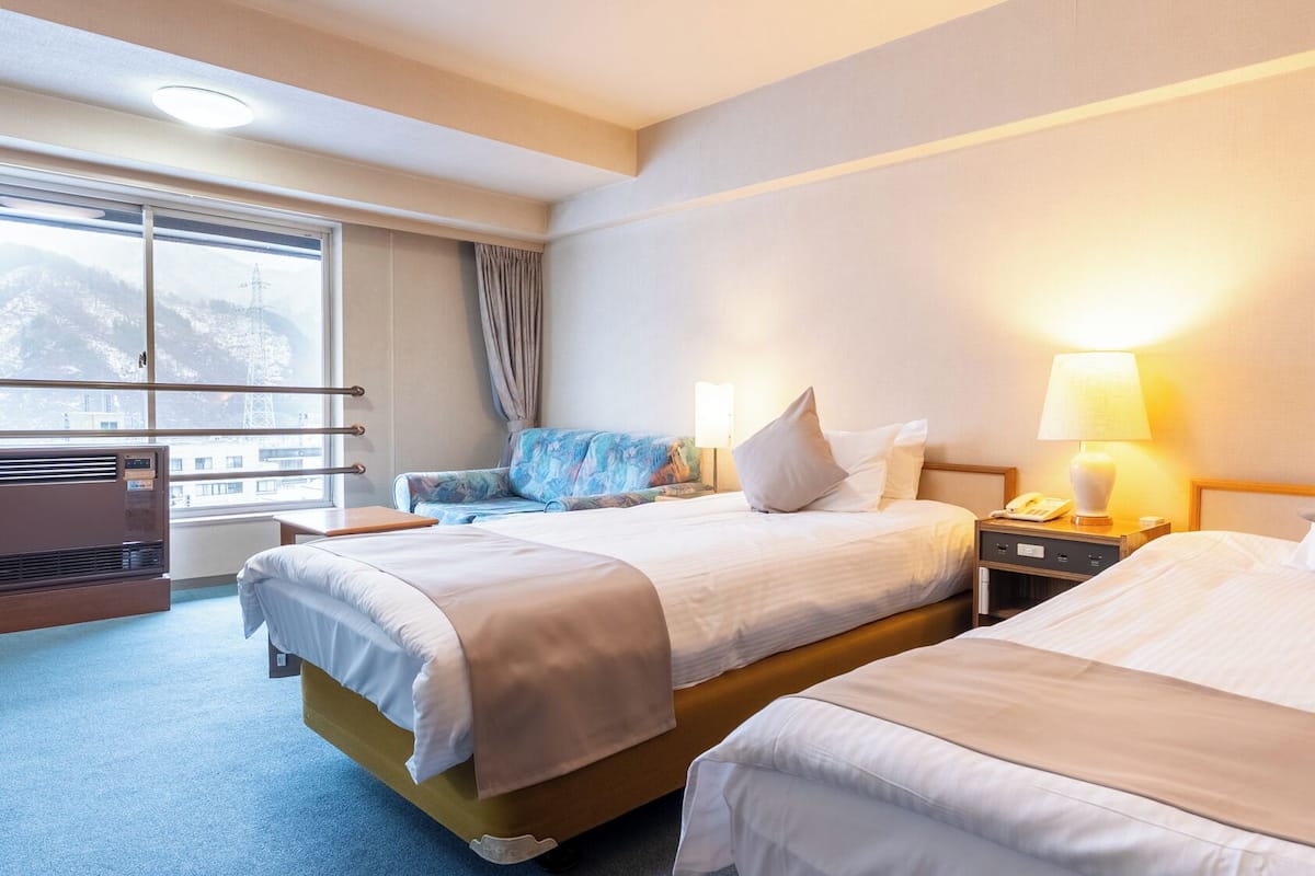 A hotel room with two beds, a desk, a chair, a window with a view of mountains, and a heater.