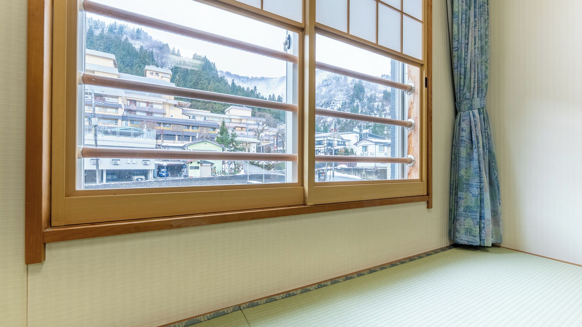 A room with a large window overlooking a residential area, a tatami mat floor, and a curtain on the right.