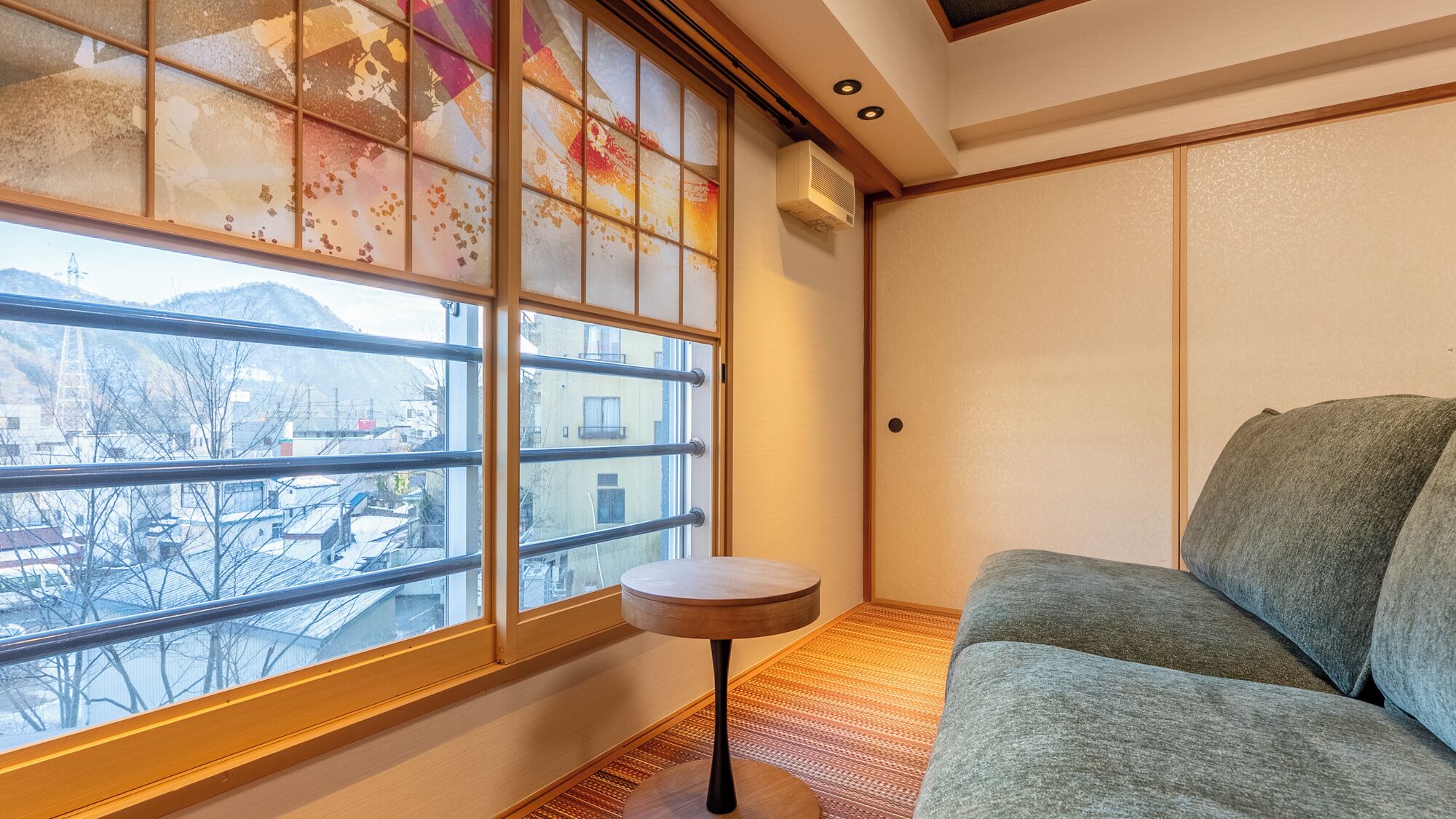 A room with a large window, a sofa, a small round table, and a wall-mounted air conditioner.