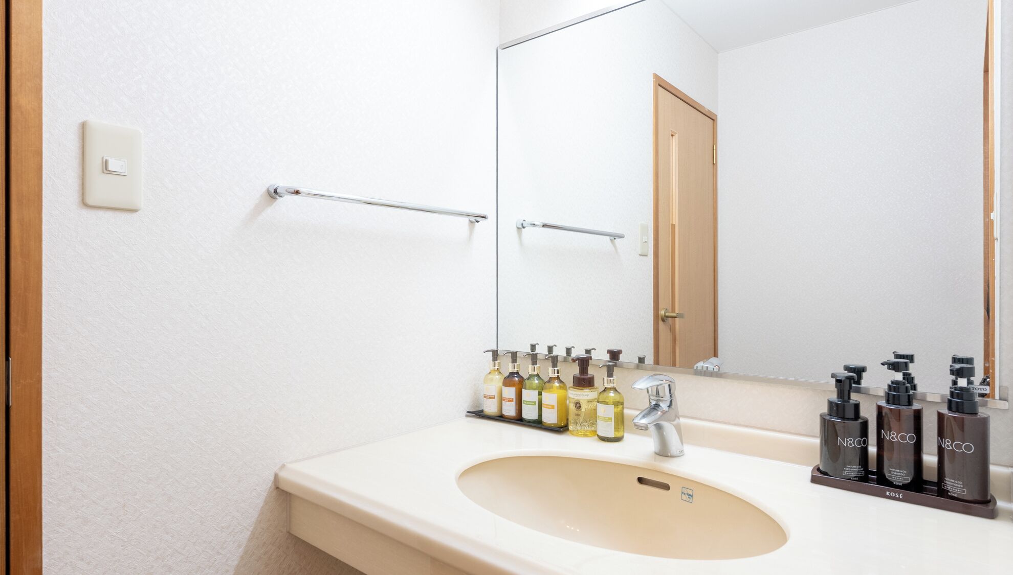 A bathroom with a beige sink, a mirror, and a handrail. There are toiletries on the sink and a door in the background.