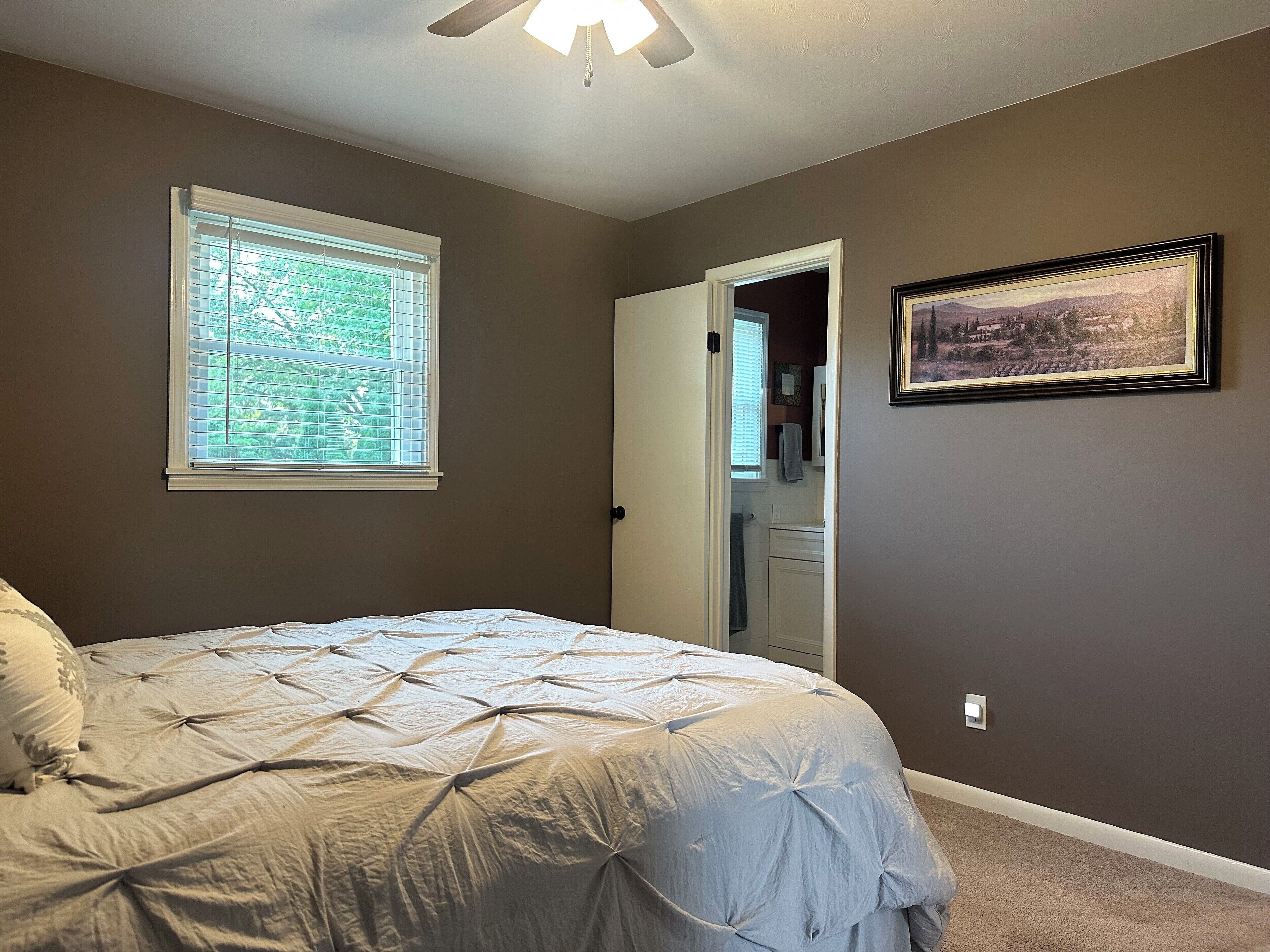 A bedroom with a bed, a window with blinds, a door leading to a bathroom, and a framed picture on the wall.