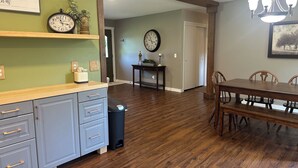 A dining area with a wooden table and chairs, a kitchen with a counter and drawers, a trash can, and a hallway with a door.