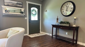 A living room with a wooden floor, a white armchair, a dark wooden console table, and a large wall clock.