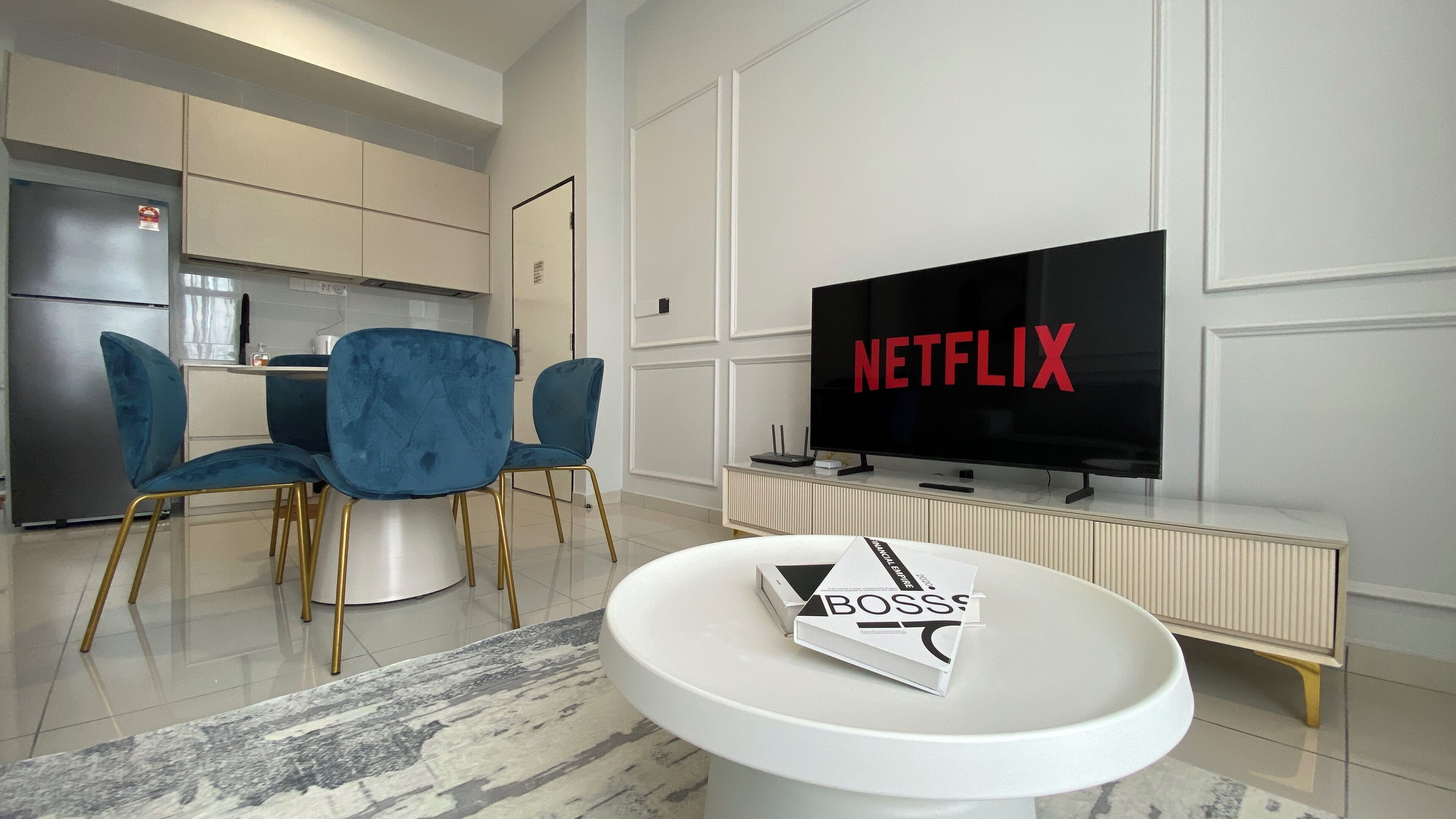 A modern living room with a flat-screen TV displaying 'NETFLIX', a dining area with blue chairs, and a white coffee table with a magazine.