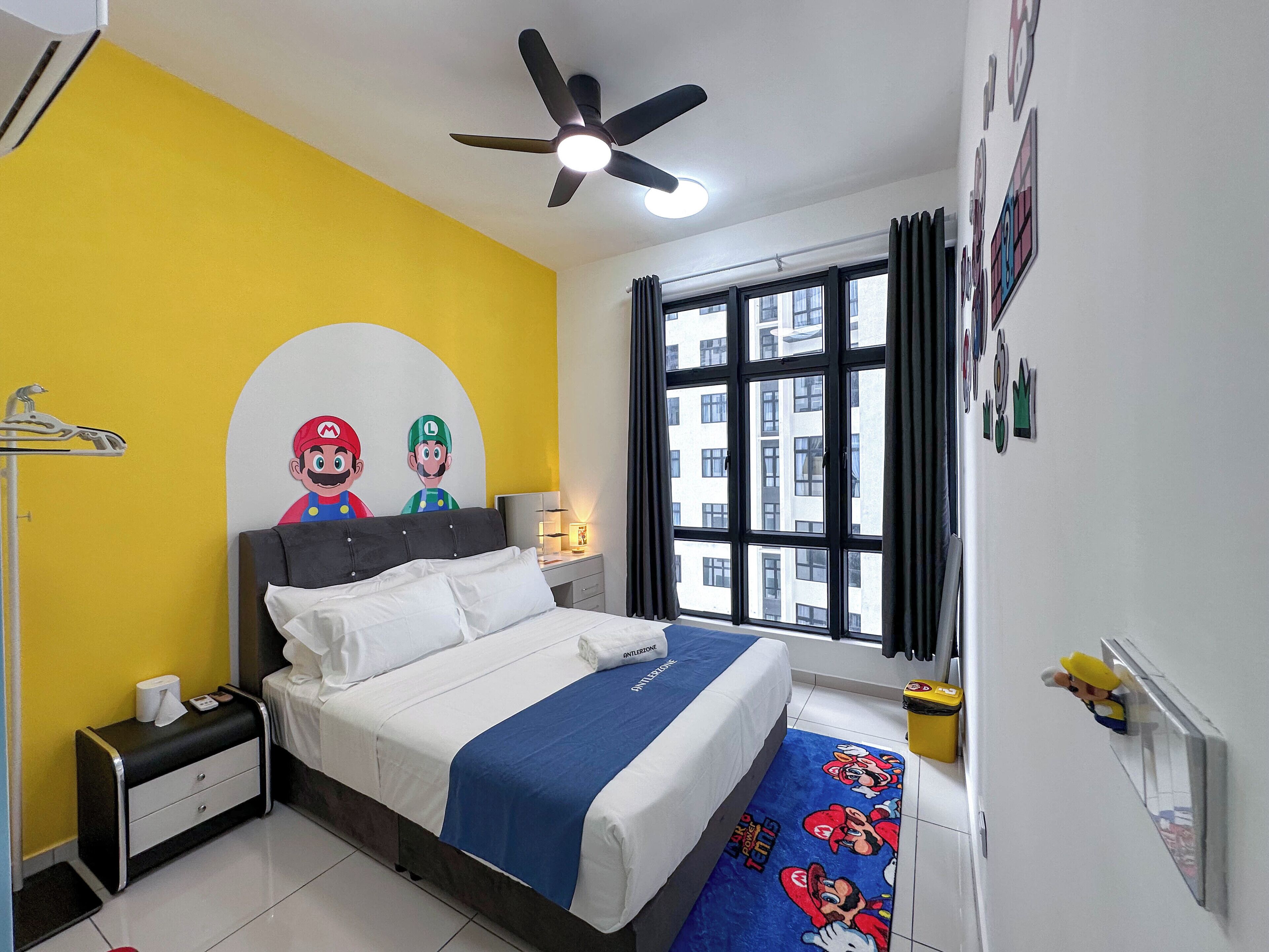 Une chambre avec un lit, une table de chevet, un ventilateur de plafond, une fenêtre avec des rideaux et un mur décoré de posters de Mario et Luigi.