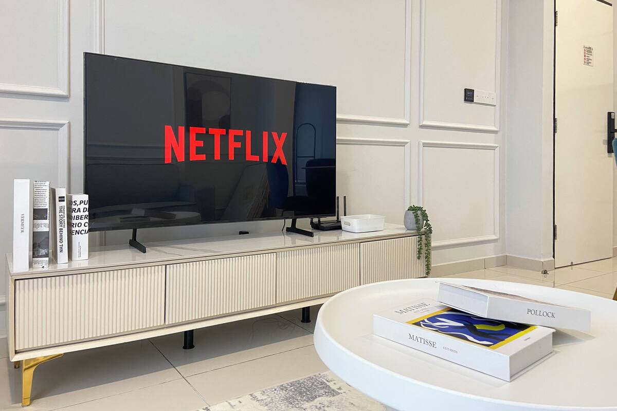 Un salon moderne doté d’un grand téléviseur à écran plat affichant le logo de Netflix, d’une console blanche sur laquelle sont posés des livres et d’une table basse ronde sur laquelle se trouvent des ouvrages d’art.