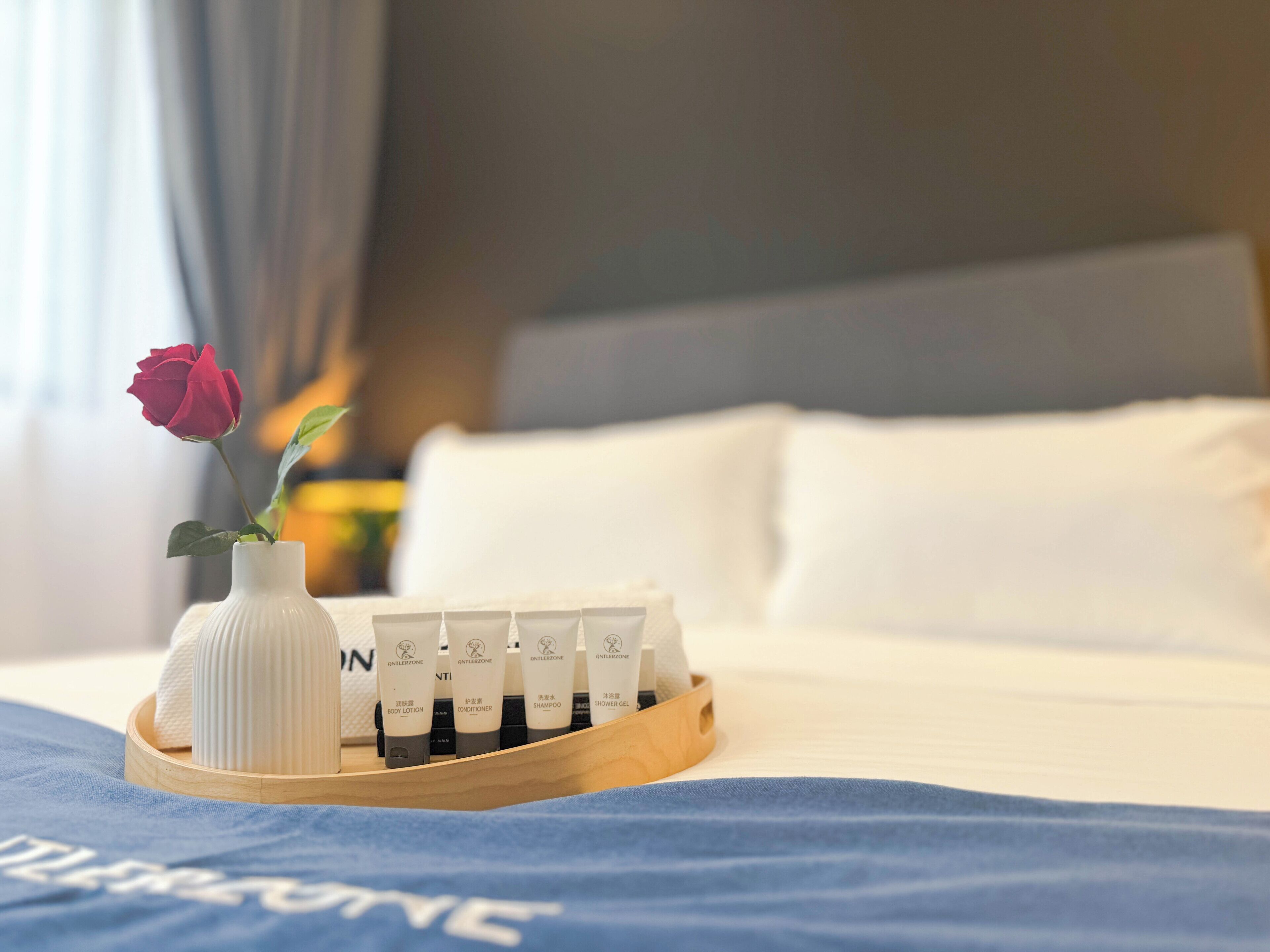 A neatly arranged tray of toiletries on a bed with a blue blanket, a white vase with a single rose, and a wooden headboard.