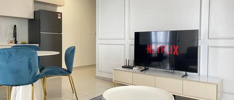 Ein modernes Wohnzimmer mit einem Flachbildfernseher, der das Netflix-Logo zeigt, einem blauen Sessel, einem weißen Couchtisch und einer Küche im Hintergrund.