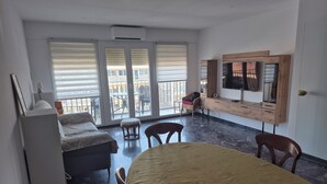 Une chambre comprenant un lit, une table à manger, des chaises, un téléviseur, un bureau et un balcon aménagé avec du mobilier d’extérieur.