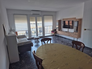Ein Zimmer mit einem Bett, einem Esstisch, Stühlen, einem Fernseher, einem Schreibtisch und einem Balkon mit Gartenmöbeln.