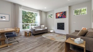 A modern living room with a fireplace, a dining area, and large windows offering a view of trees. - Perch - Modern End-Unit Surrounded by Nature (Oak Harbor)