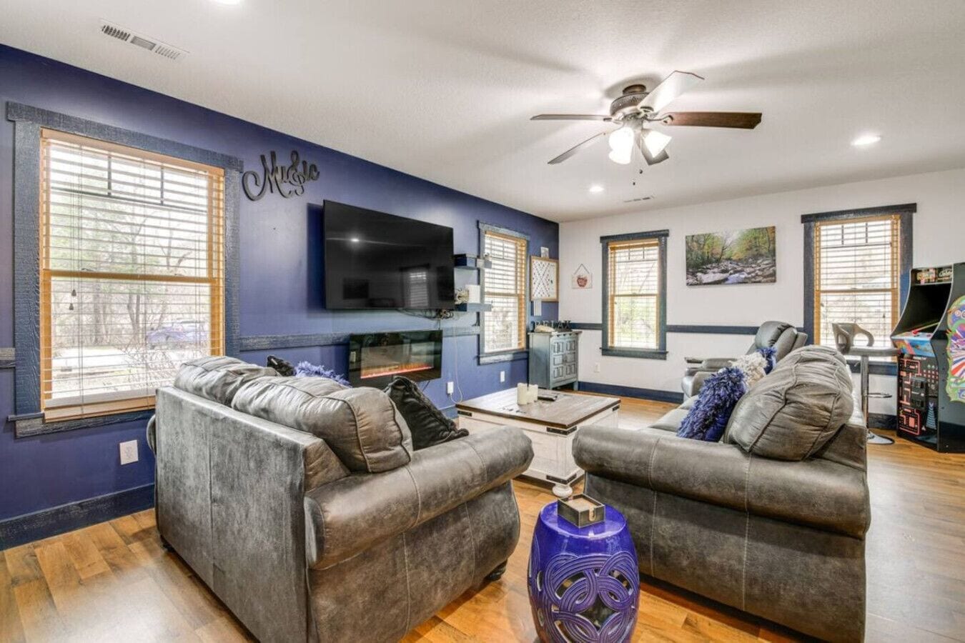 A living room with two grey sofas, a flat-screen TV, a ceiling fan, and a large window with blinds.