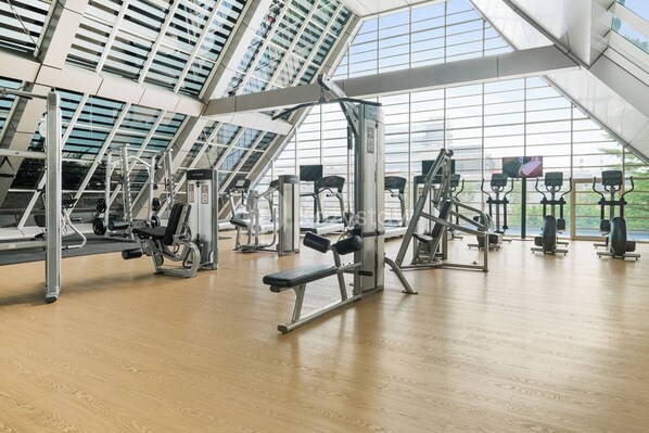 A modern gym with various exercise machines, large windows, and a wooden floor.