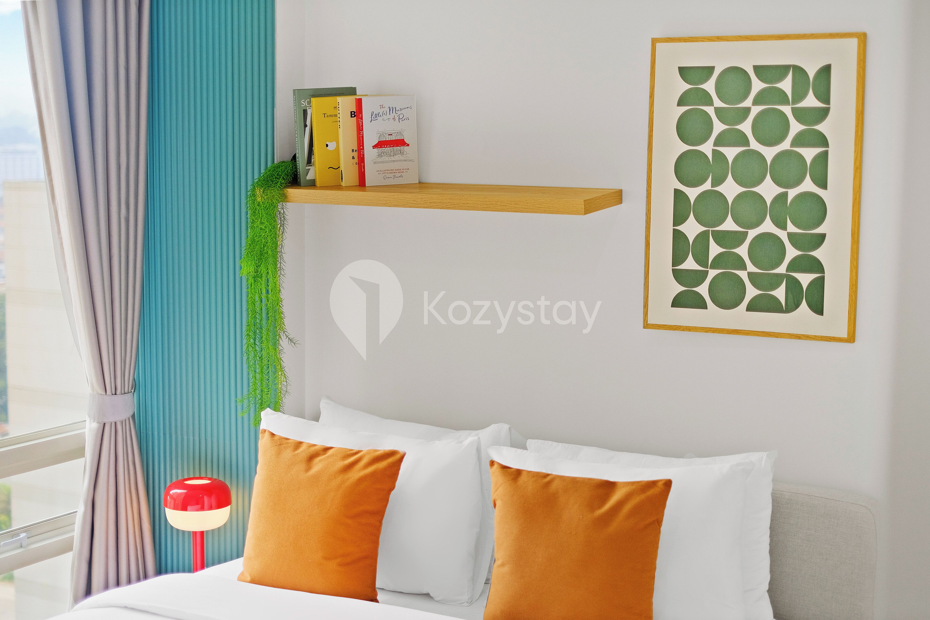 A hotel room with a bed, pillows, a shelf with books, a framed artwork, and a plant.