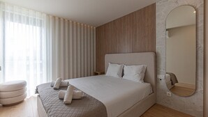 A modern bedroom with a large bed, a wooden headboard, a round mirror, and a view of the city through sheer curtains. - GuestReady - Elegant beachside stay in Apúlia (Apúlia e Fão)
