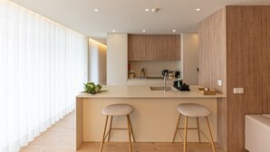 A modern kitchen with a central island, two bar stools, wooden cabinets, and a large window with sheer curtains. - GuestReady - Elegant beachside stay in Apúlia (Apúlia e Fão)