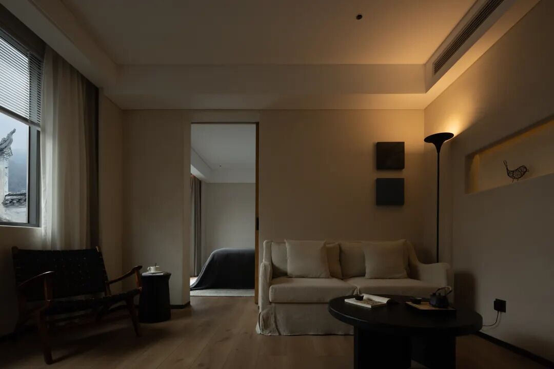 A modern living room with a sofa, a small table, and a chair. There is a window with blinds, a lamp, and a picture frame on the wall.