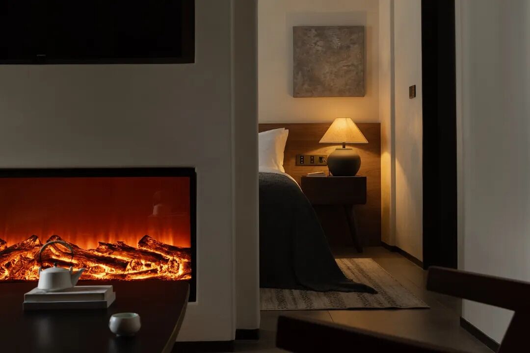 A cozy hotel room with a fireplace, a bed with a nightstand, a lamp, and a painting on the wall.