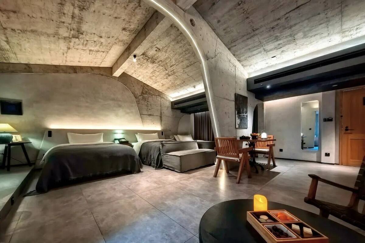 A modern, compact bedroom with a curved ceiling, a sofa bed, a dining area with a table and chairs, and a small side table with a candle.