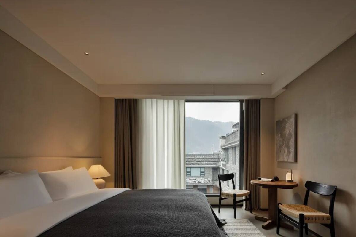 A hotel room with a bed, a desk, a chair, and a large window offering a view of buildings and mountains.