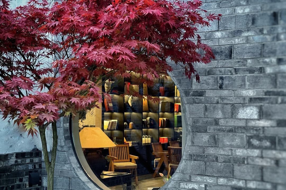 A circular window with a view of a room containing a bookshelf, a lamp, and a chair.