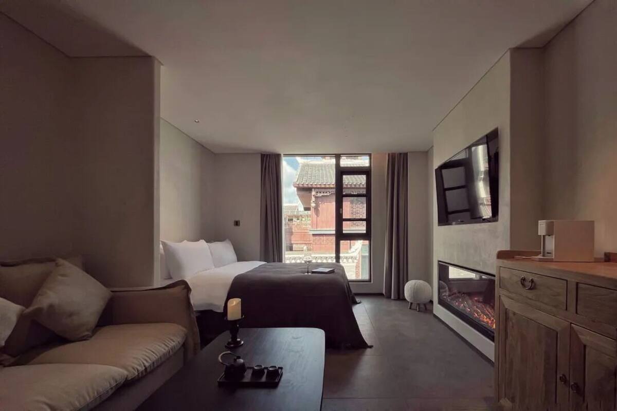 A modern hotel room with a bed, a sofa, a TV, a fireplace, and a view of buildings outside.