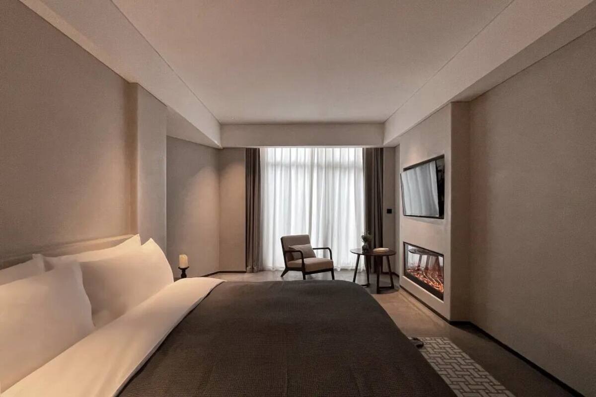 A modern hotel room with a large bed, a seating area with a chair and a small table, a fireplace, and a large window with curtains.