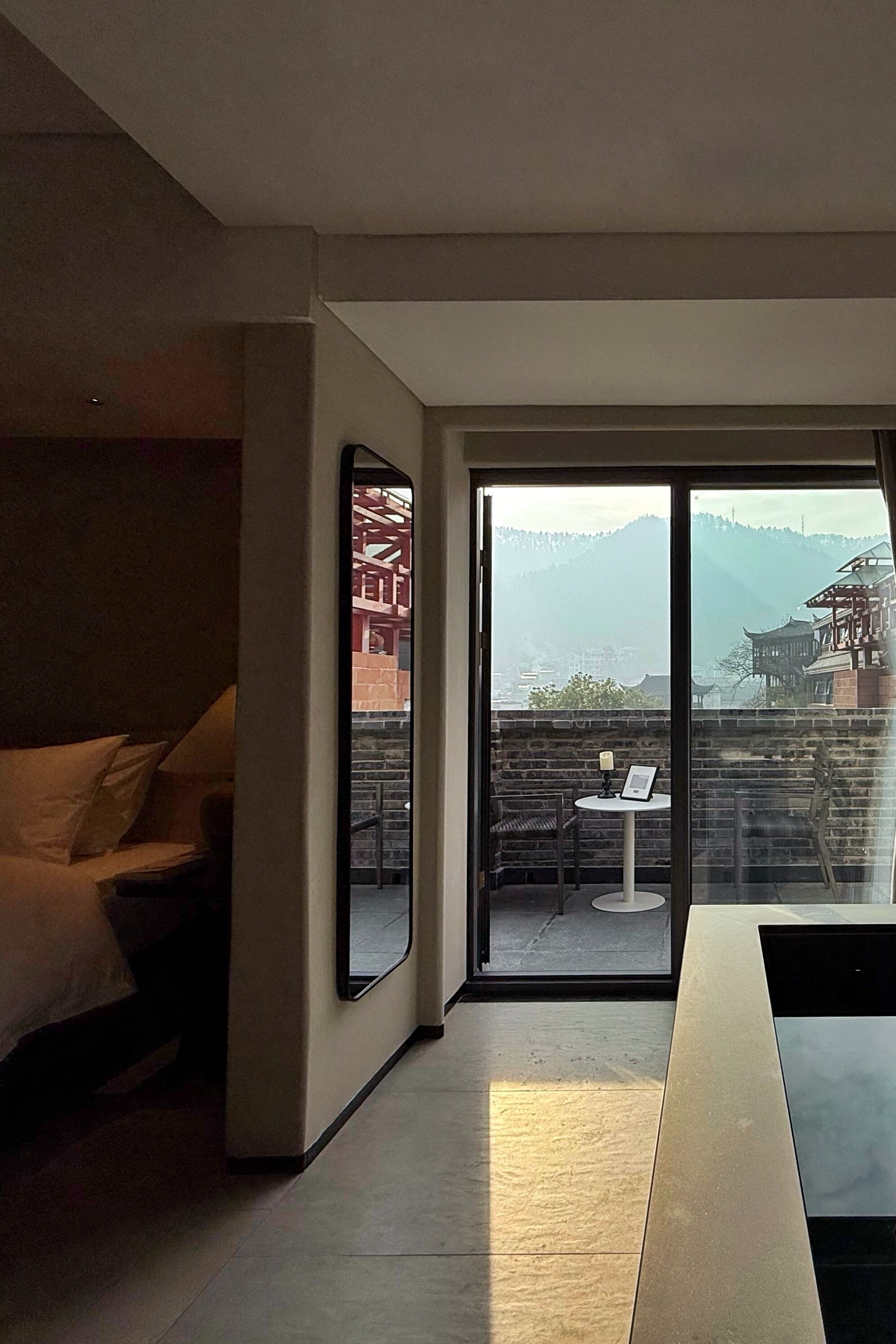 A modern hotel room with a large sliding glass door leading to a balcony. The room features a bed, a small table with a chair, and a large mirror.