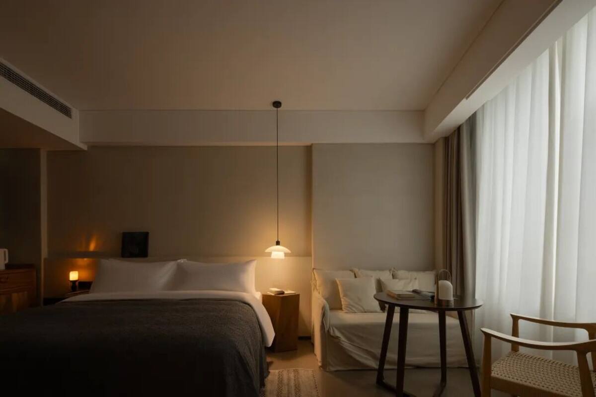 A hotel room with a bed, bedside table, lamp, chair, and a view of the outside through large windows.