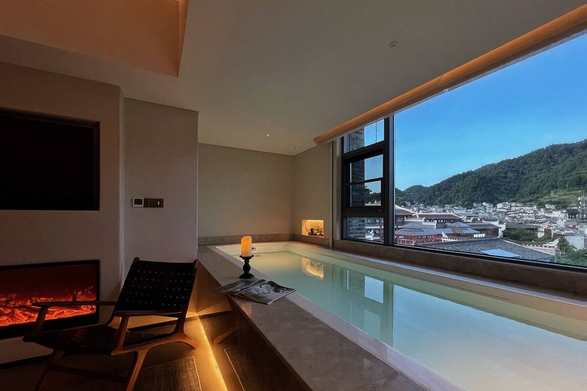 A modern room with a large window overlooking a cityscape, a fireplace, and a pool area.