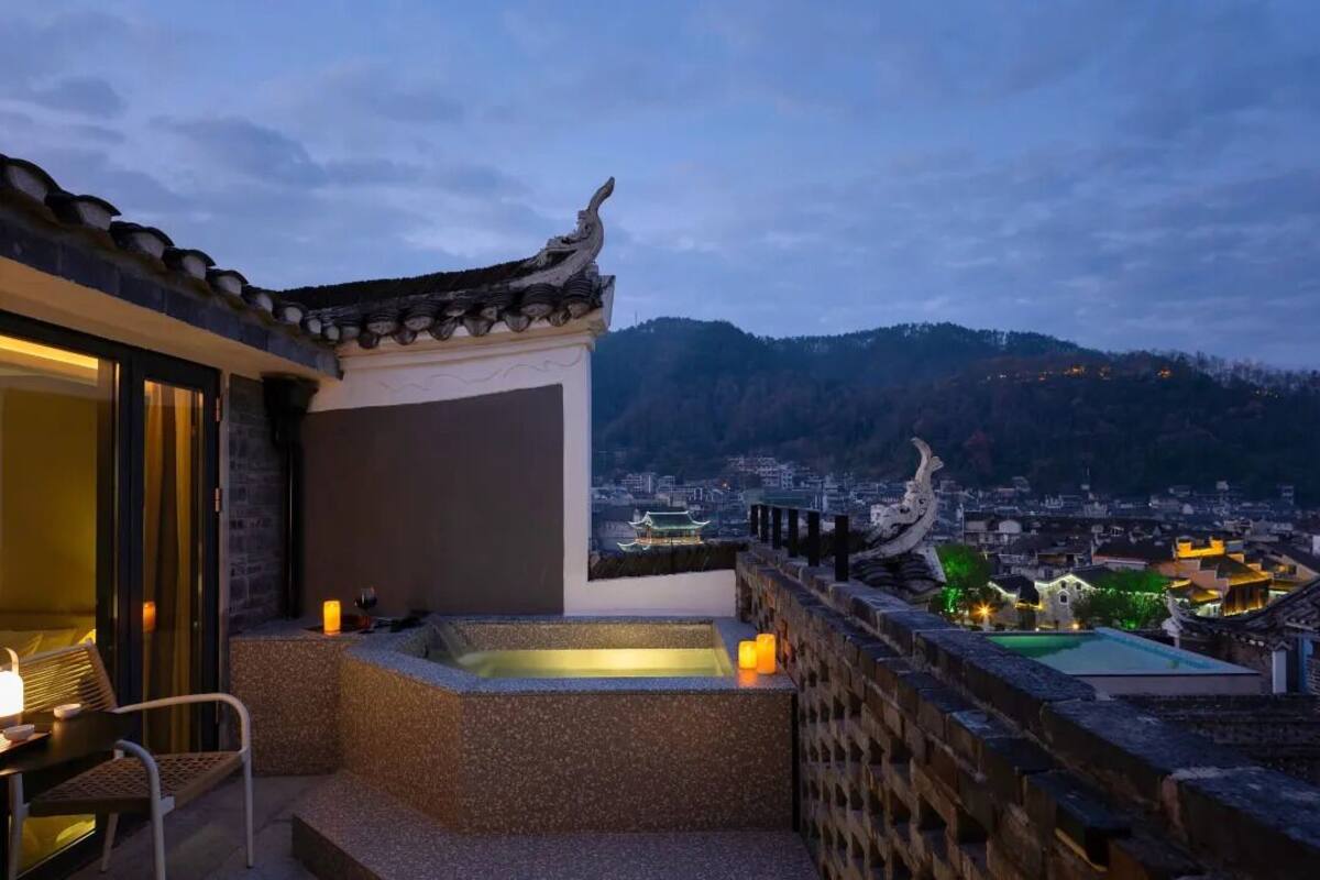 A rooftop terrace with a traditional Chinese architectural style, a hot tub, and a view of a cityscape and mountains in the background.