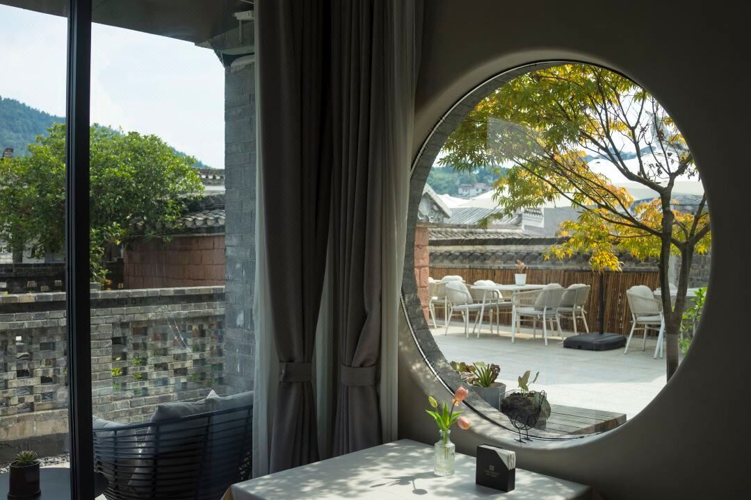 A room with a large round window offering a view of traditional architecture and a patio area with chairs and tables.