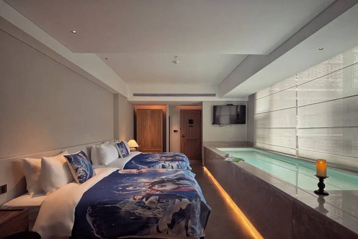 A hotel room with a bed, a small bedside table, a TV mounted on the wall, and a window with a candle on the sill.