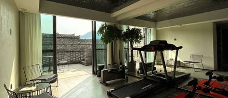A modern living room with a treadmill, a dining area with chairs, a large window with a view of traditional architecture, and a red patterned rug.