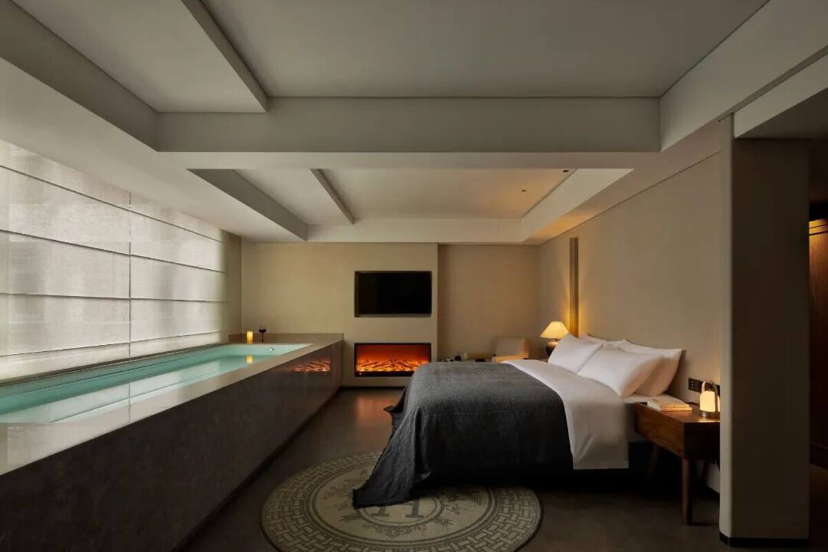 A modern hotel room with a large bed, a fireplace, a glass bathtub, and a flat-screen TV.
