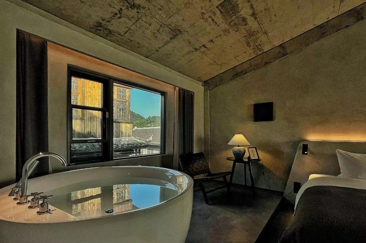 A hotel room with a large bathtub, a bed, a desk with a lamp, and a view of a building through the window.