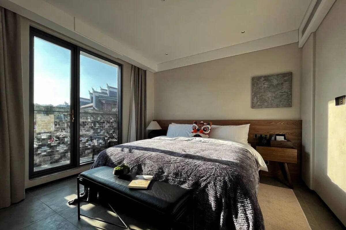 A modern hotel room with a large bed, a bench, a nightstand, and a view of a cityscape through floor-to-ceiling windows.
